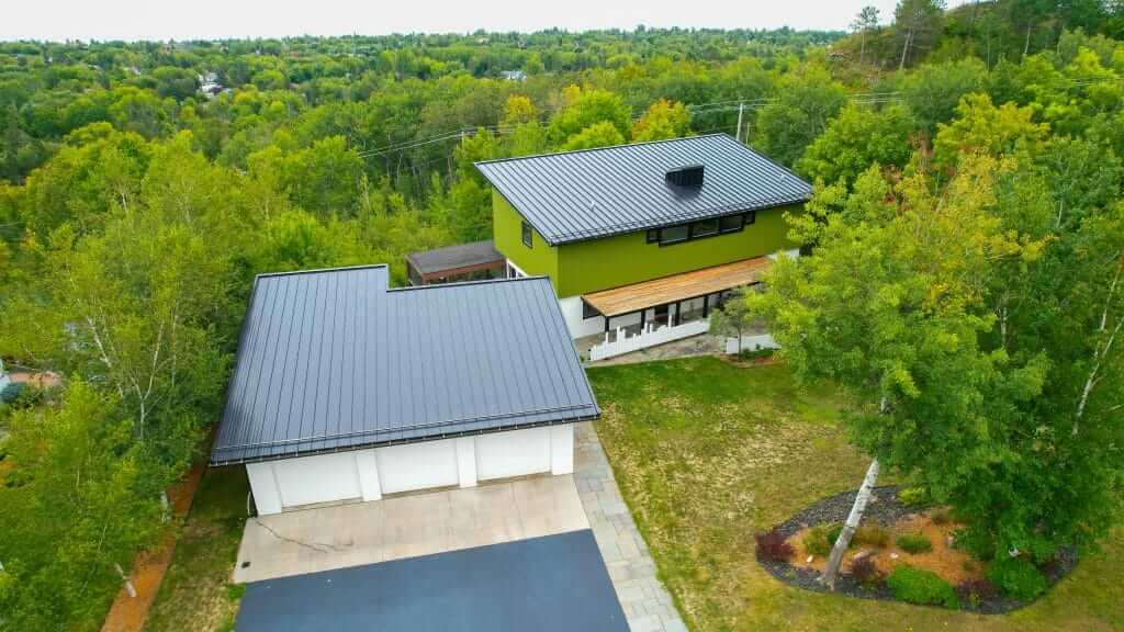 Modern house with a black metal roof and green siding, surrounded by lush trees, showcasing expert roofing installation by Perrault Construction in Two Harbors, MN.