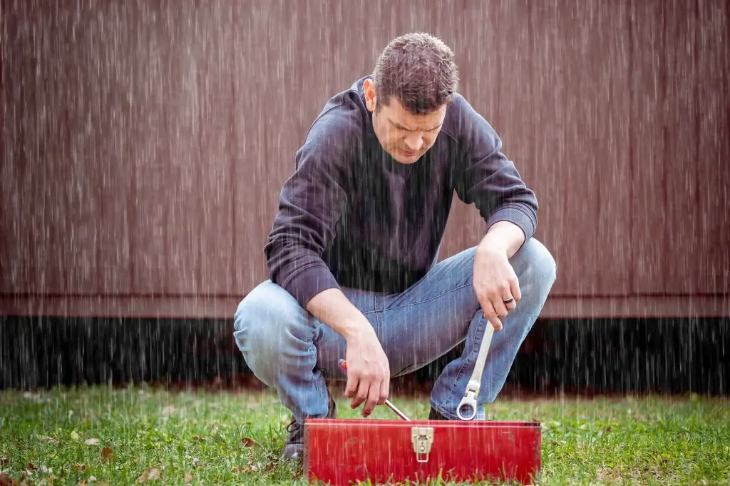 Man crouching in the rain, examining tools in a red toolbox, highlighting challenges of roofing work in wet conditions.